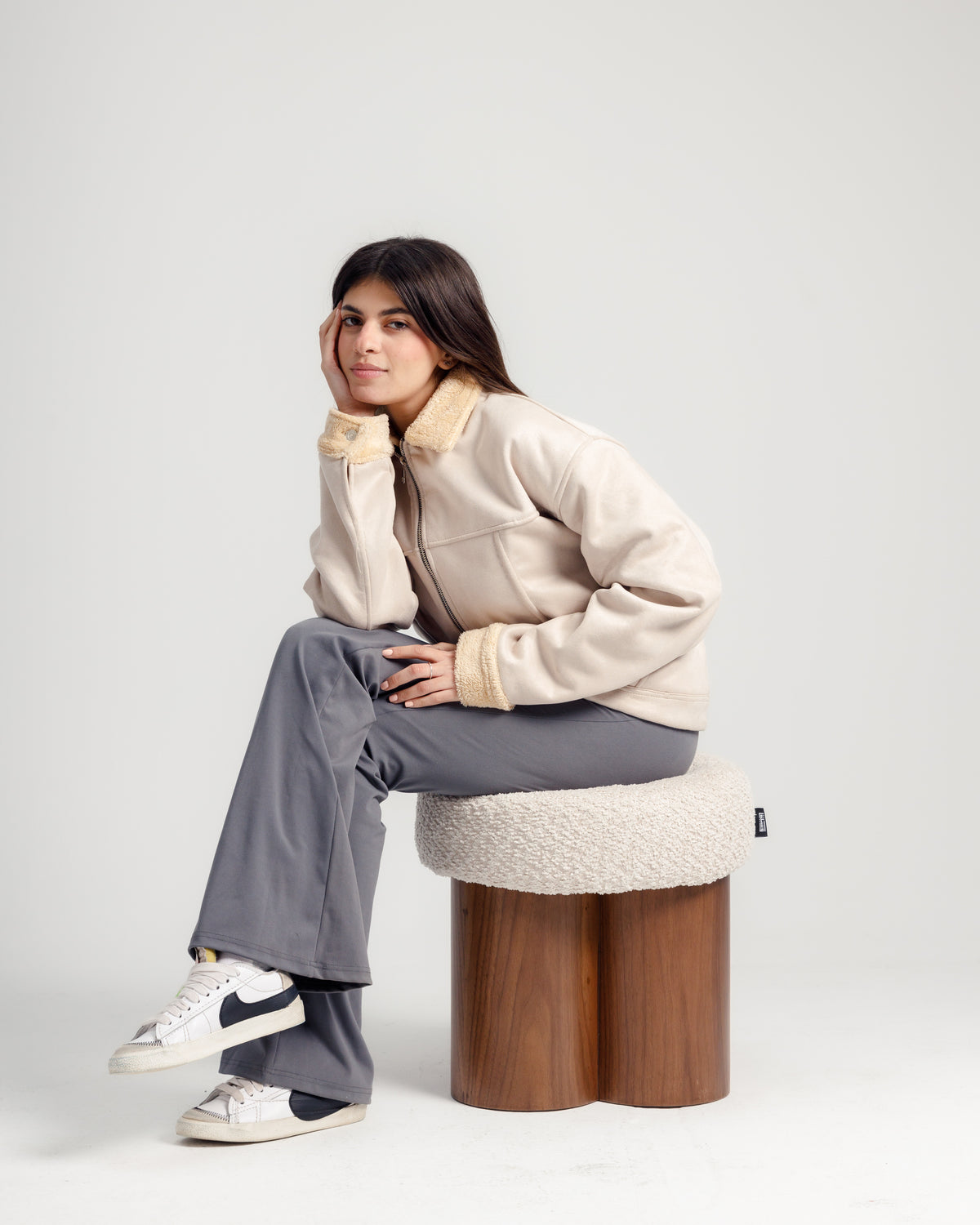A young woman sits on a stylish pouf with a soft cream cushion and a wooden base, wearing a beige jacket with a fur collar, gray pants, and white sneakers. She rests her chin on her hand, exuding a relaxed and casual vibe.