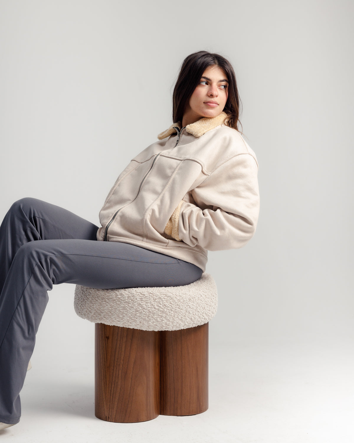 A young woman sits comfortably on a round pouf with a soft cream cushion and a walnut wood base, wearing a beige zip-up jacket and gray pants. She poses with one hand in her pocket, exuding a relaxed and casual vibe.