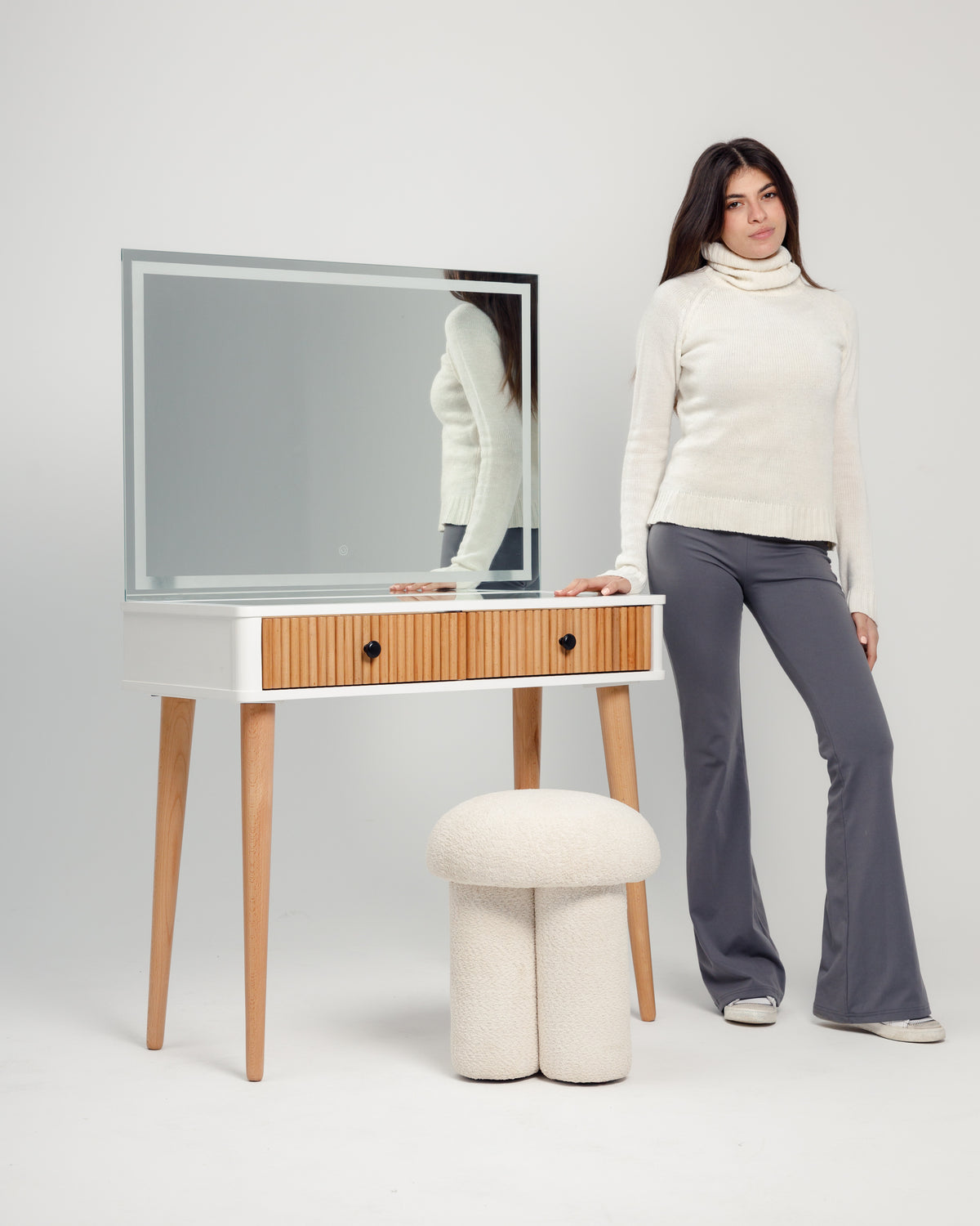 A woman stands beside a modern vanity table with a large mirror, wearing a cozy white sweater and gray pants. The table features a wooden front and two drawers, while a plush, off-white pouf sits on the floor next to it.