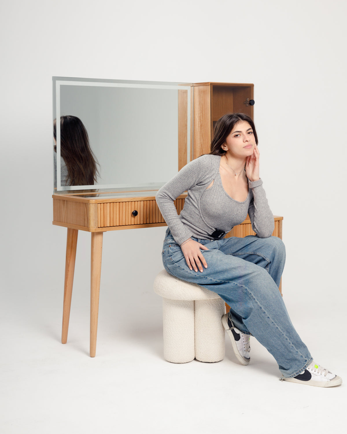 A young woman sits casually on a textured off-white pouf, wearing a gray long-sleeve top and baggy blue jeans. She poses thoughtfully beside a wooden vanity with a large mirror, set against a simple white backdrop.