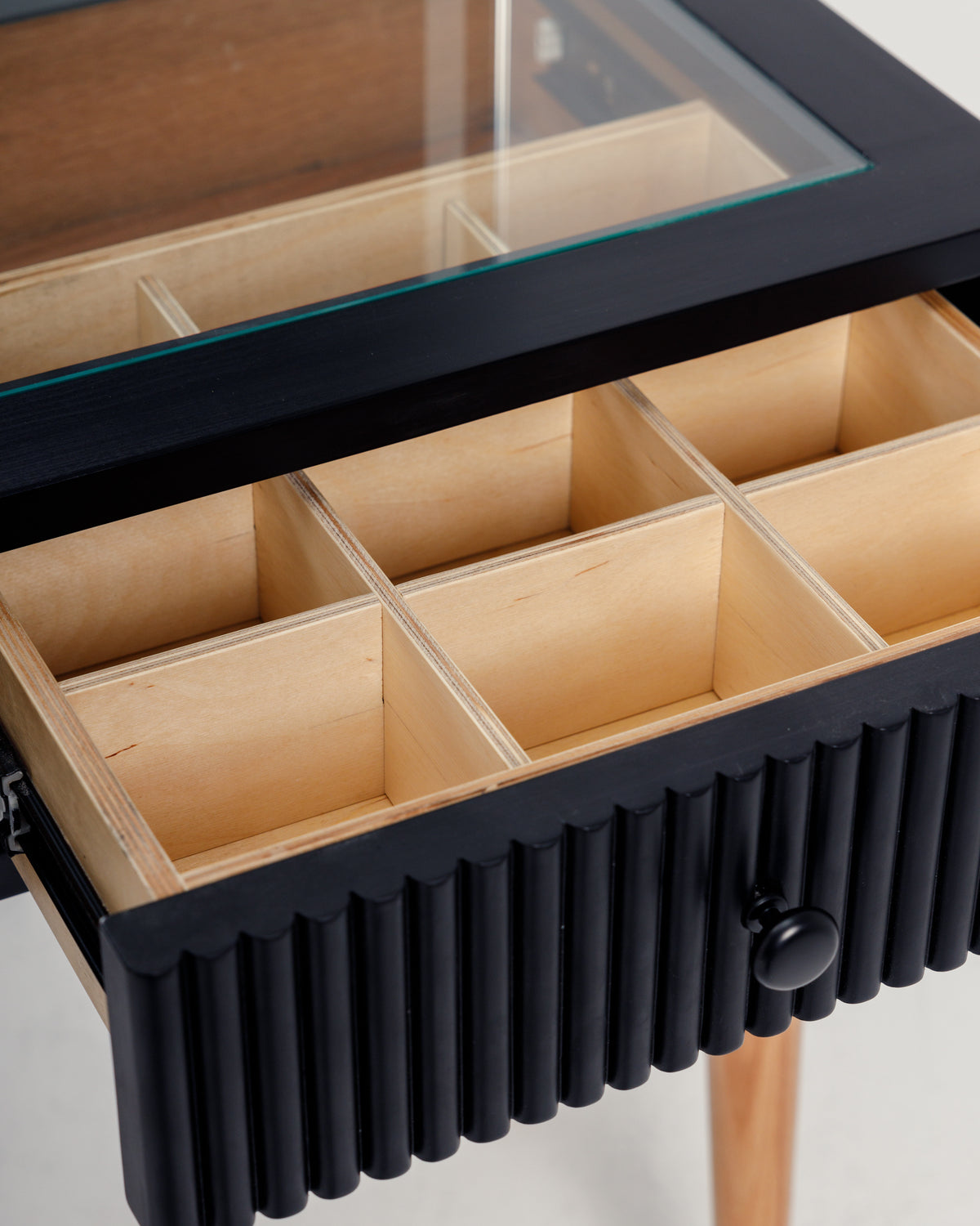 A close-up of a stylish black dresser featuring a glass top and a ribbed drawer front. The drawer is open, revealing multiple compartments made of light wood, ideal for organizing various items.