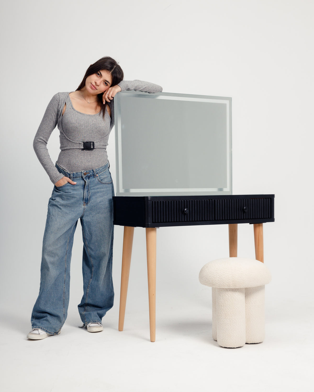 A young woman leans against a modern black dresser with a large mirror, wearing a fitted gray top and baggy blue jeans. Beside the dresser, a stylish, round stool adds a touch of contemporary design to the setting.