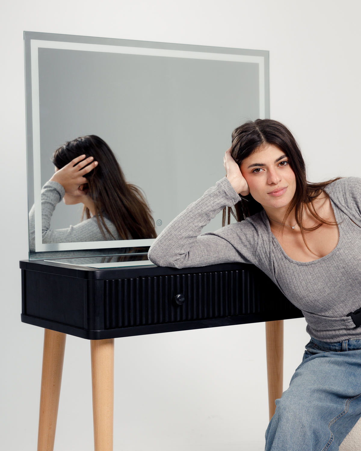 A young woman with long dark hair leans casually against a sleek black dresser with a large mirror. She wears a light gray long-sleeve top and a denim skirt, showcasing a modern and stylish look in a minimalist setting.
