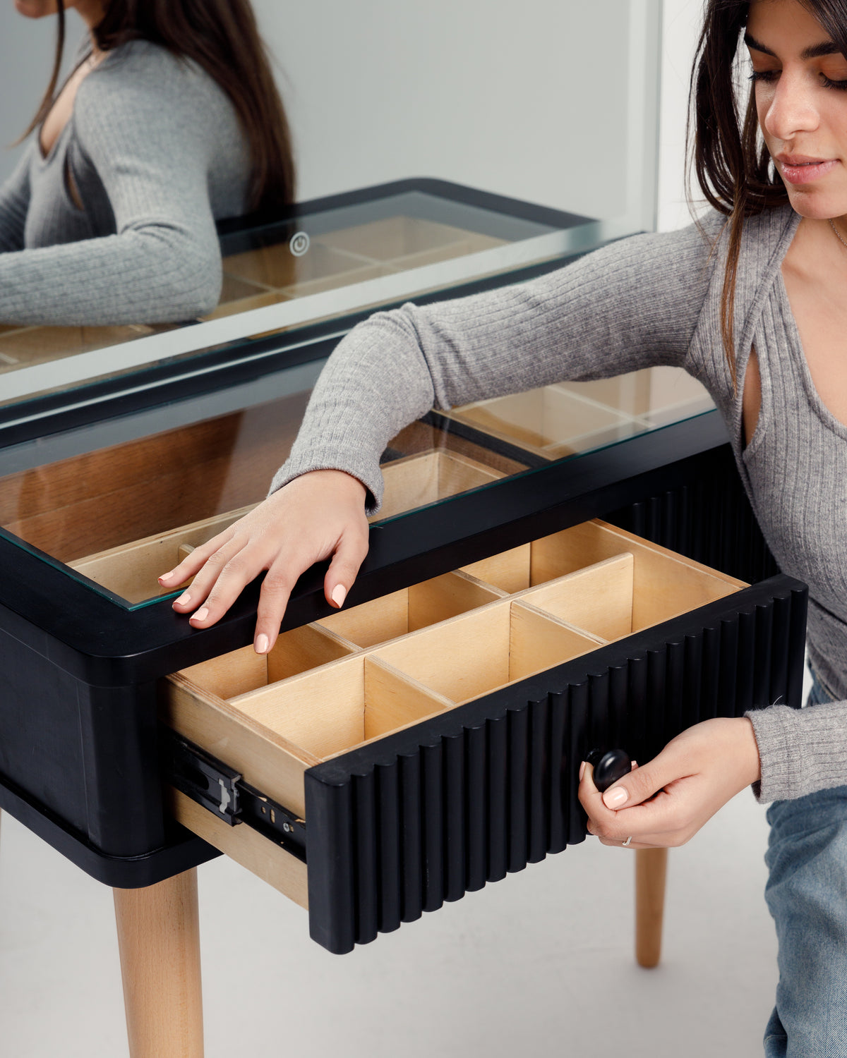 A woman in a gray sweater is opening a drawer of a modern black dresser with a glass top and wooden legs. The interior of the drawer features organized compartments, and a large mirror is visible in the background.