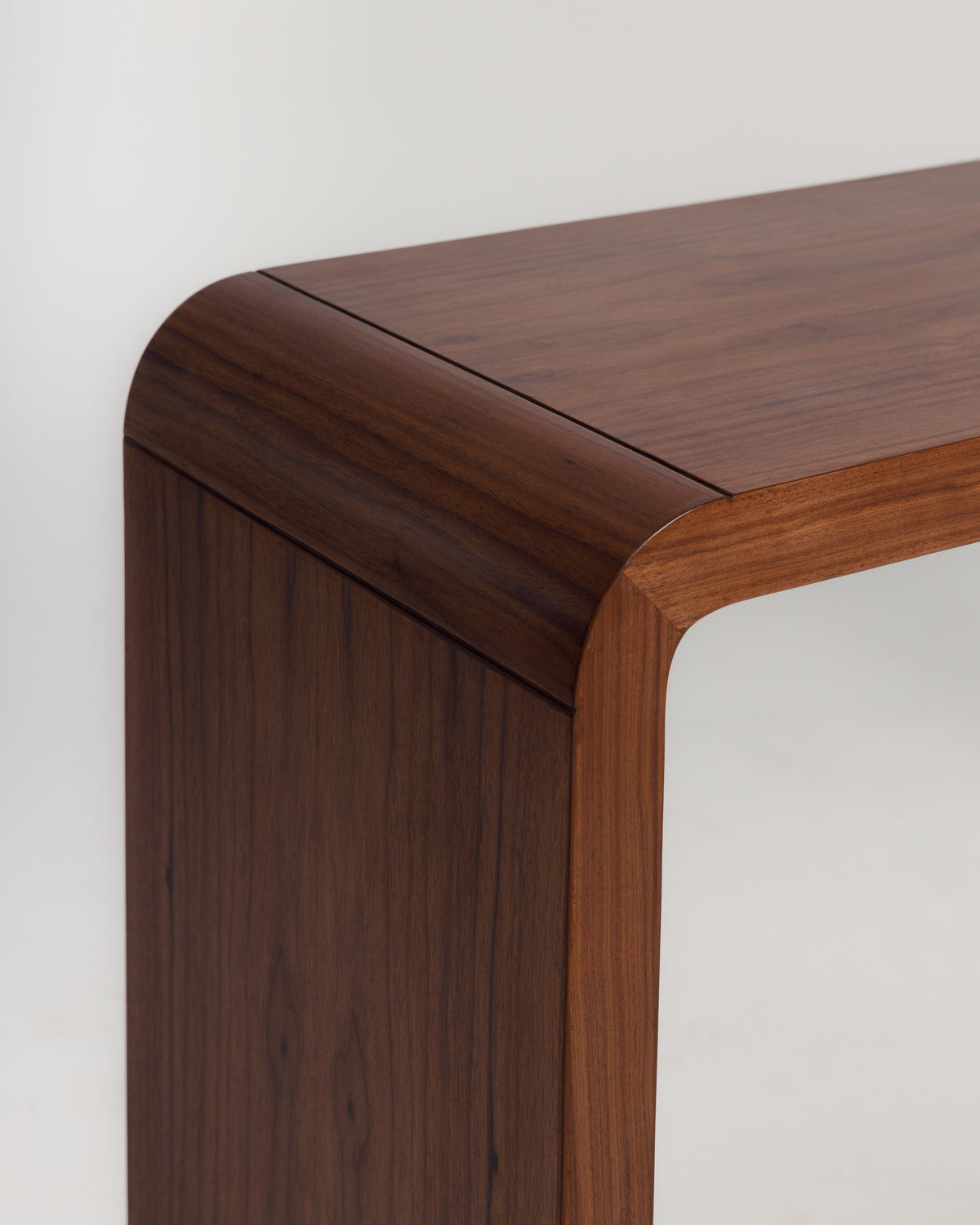 A close-up view of a walnut wood console table, showcasing its smooth, rounded edges and rich brown grain. The handcrafted finish highlights the natural beauty of the wood, making it an elegant addition to any space.