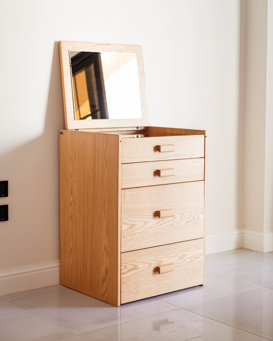 A wooden petite dresser with a natural finish features four drawers and an open mirror on top. It is positioned against a light-colored wall, showcasing its sleek design and space-saving functionality.