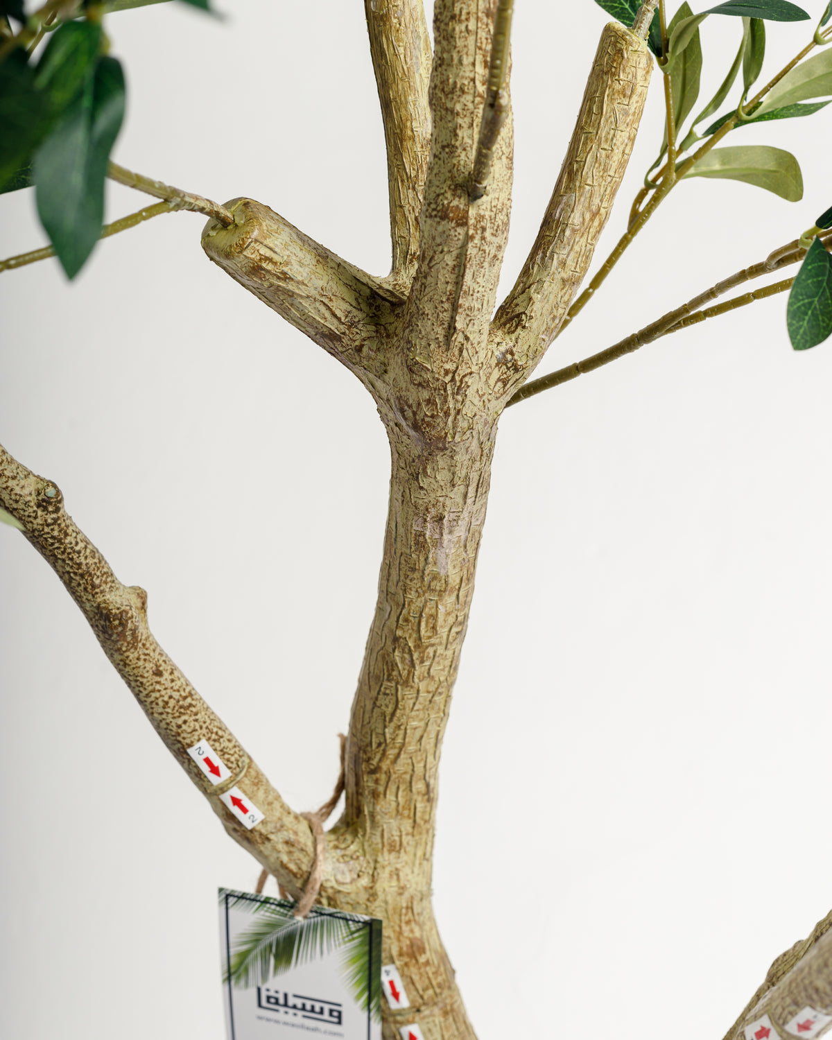 Close-up view of the textured trunk of an artificial olive tree, showcasing its realistic bark patterns and branches. Green leaves extend from the branches, highlighting the lifelike appearance of this decorative piece.