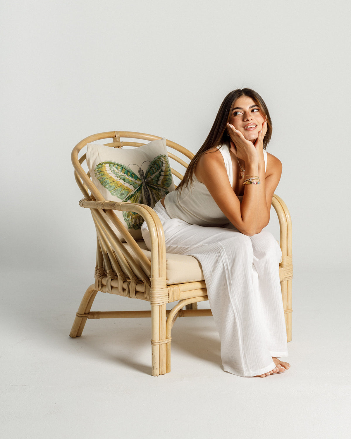 A woman sitting gracefully on a stylish bamboo chair, wearing a white outfit, with her chin resting on her hand and a thoughtful expression. The chair features a decorative cushion with a butterfly design, set against a neutral background.