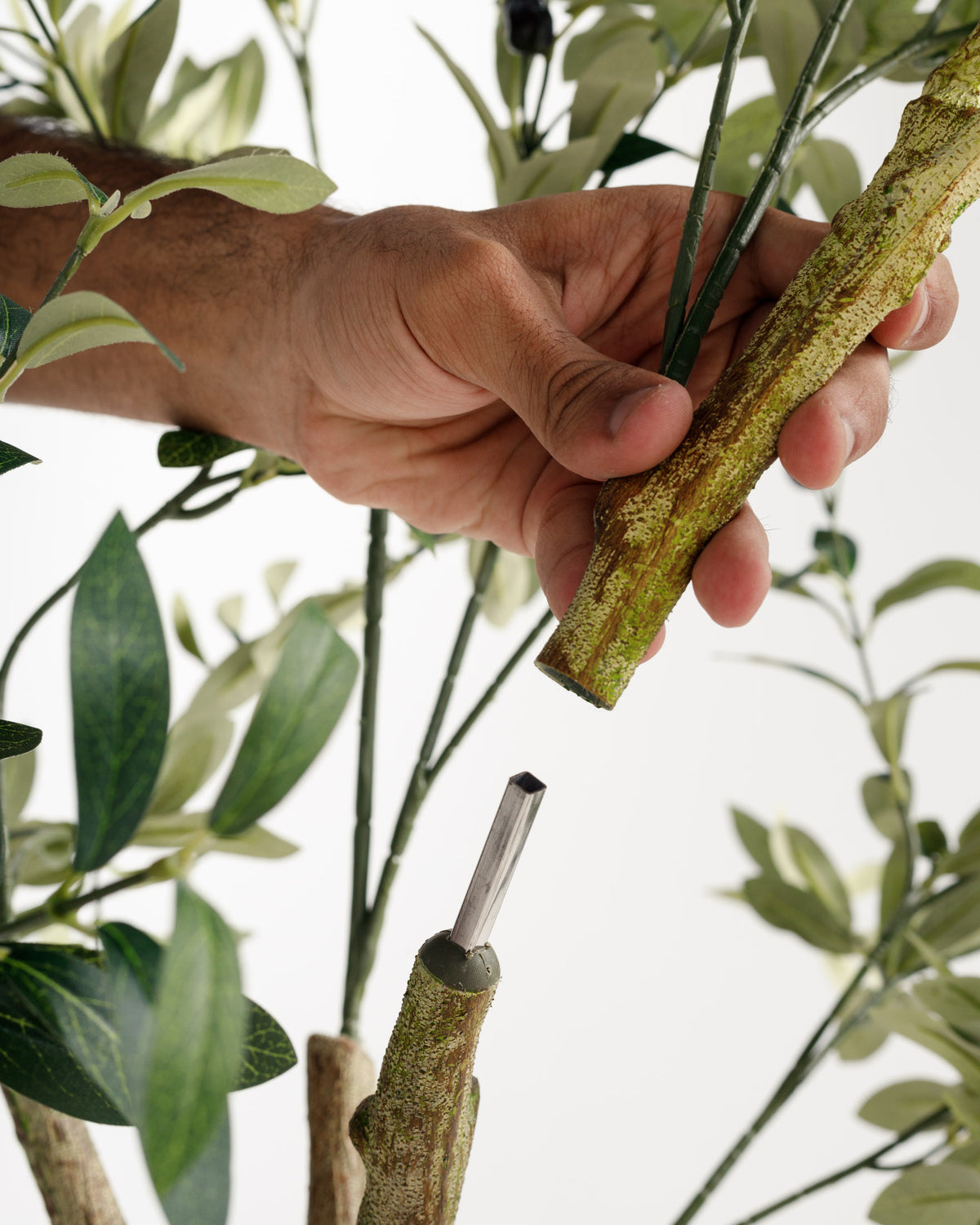 A close-up view of a persons hand connecting two segments of an artificial olive tree. The hand is holding a textured branch while green leaves and stems surround the scene, emphasizing the lifelike appearance of the plant.