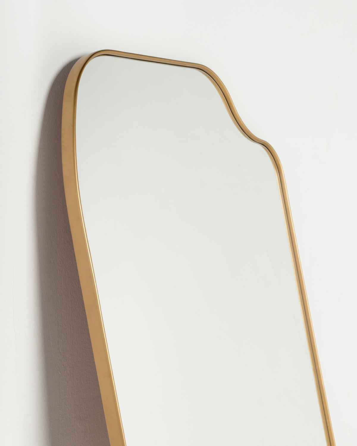 A stylish full-length mirror with a smooth, reflective surface and a gold-colored frame featuring a gently curved top edge. The mirror is mounted against a plain white wall, highlighting its elegant design.
