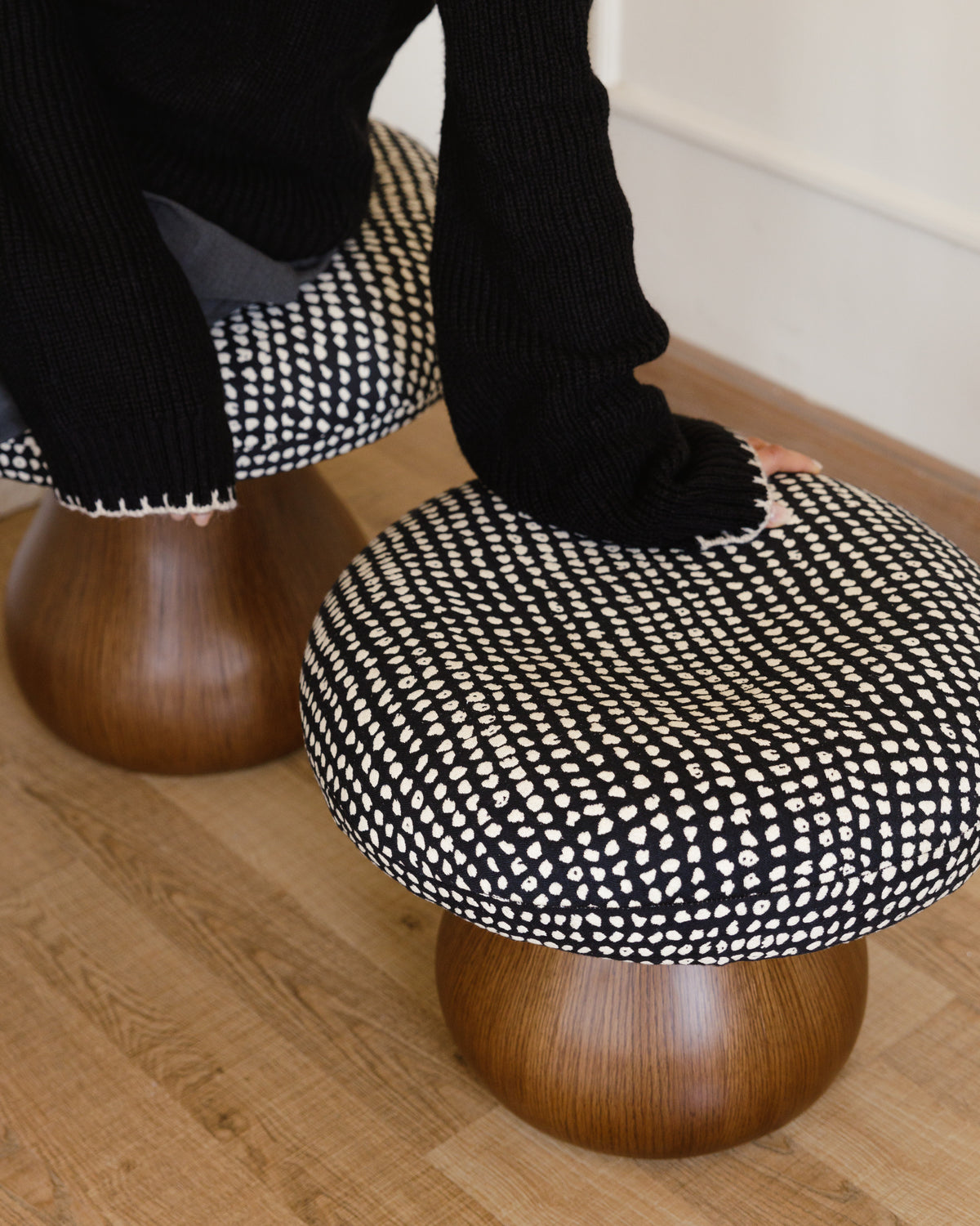 Two mushroom-shaped poufs are positioned on a wooden floor, featuring a curvaceous wooden base and a round, padded cushion with a black and white dotted pattern. A person in a black sweater is gently resting their hand on one of the poufs.