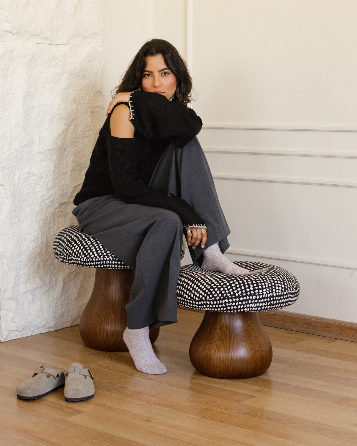 A woman with long, dark hair sits on a stylish mushroom-shaped pouf, wearing a black sweater and gray pants. The pouf features a patterned cushion atop a sleek wooden base, with a pair of shoes resting on the wooden floor nearby.