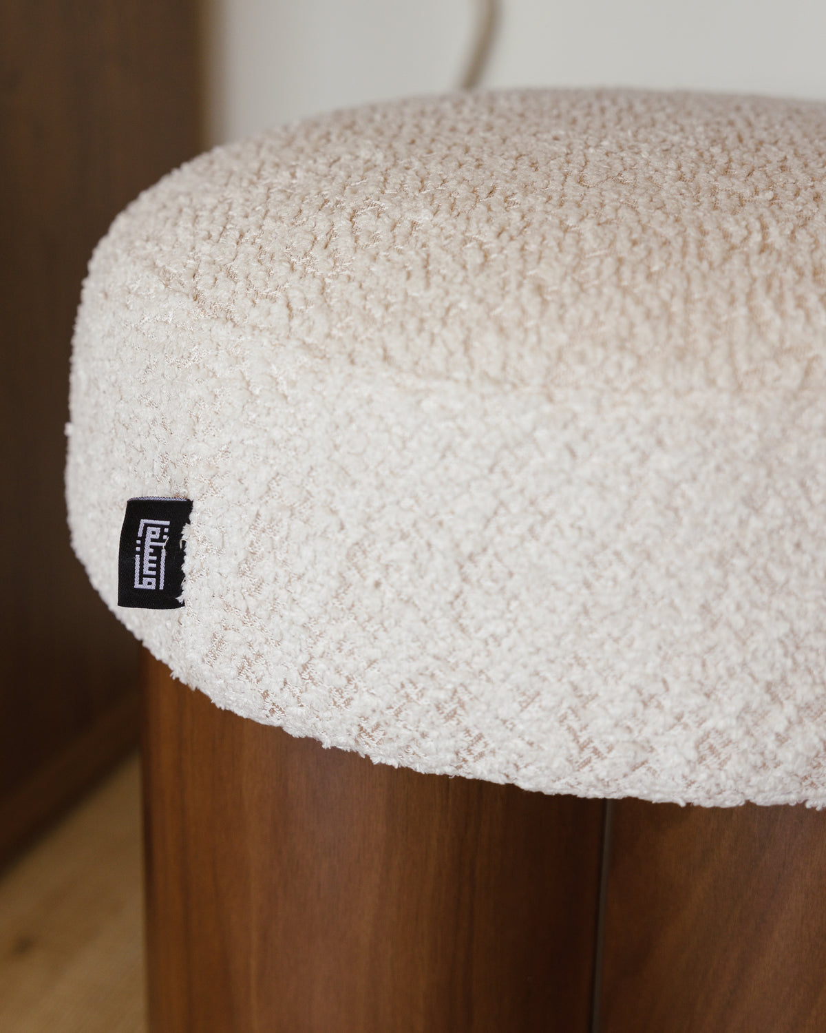 A close-up view of a cream-colored, textured pouf cushion resting on a wooden base with a unique cylindrical design. The cushion features a small black label on its side, emphasizing its stylish and cozy appeal.