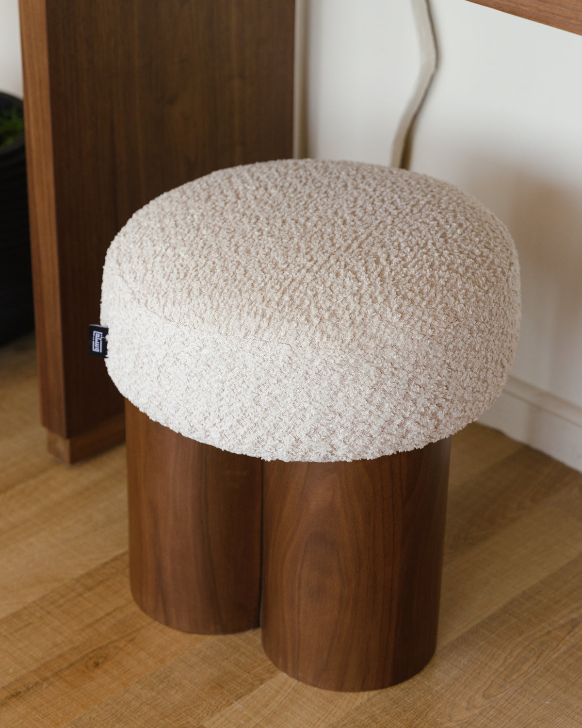 A round pouf features a cozy cream cushion with a textured surface, resting on a unique triple-cylinder walnut wood base. The design combines natural warmth and soft elegance, making it a versatile accent piece for any space.