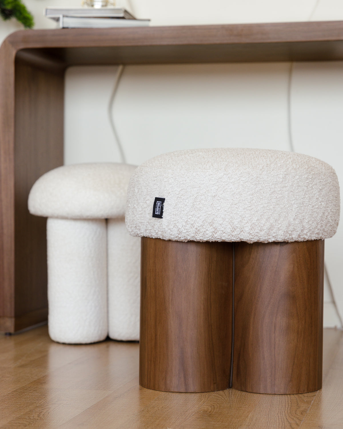 A cozy Cloud Double Pouf features a dual cylindrical wooden base with a plush, off-white bouclé cushion on top. The pouf is positioned near a wooden desk, adding a modern and stylish accent to the space.