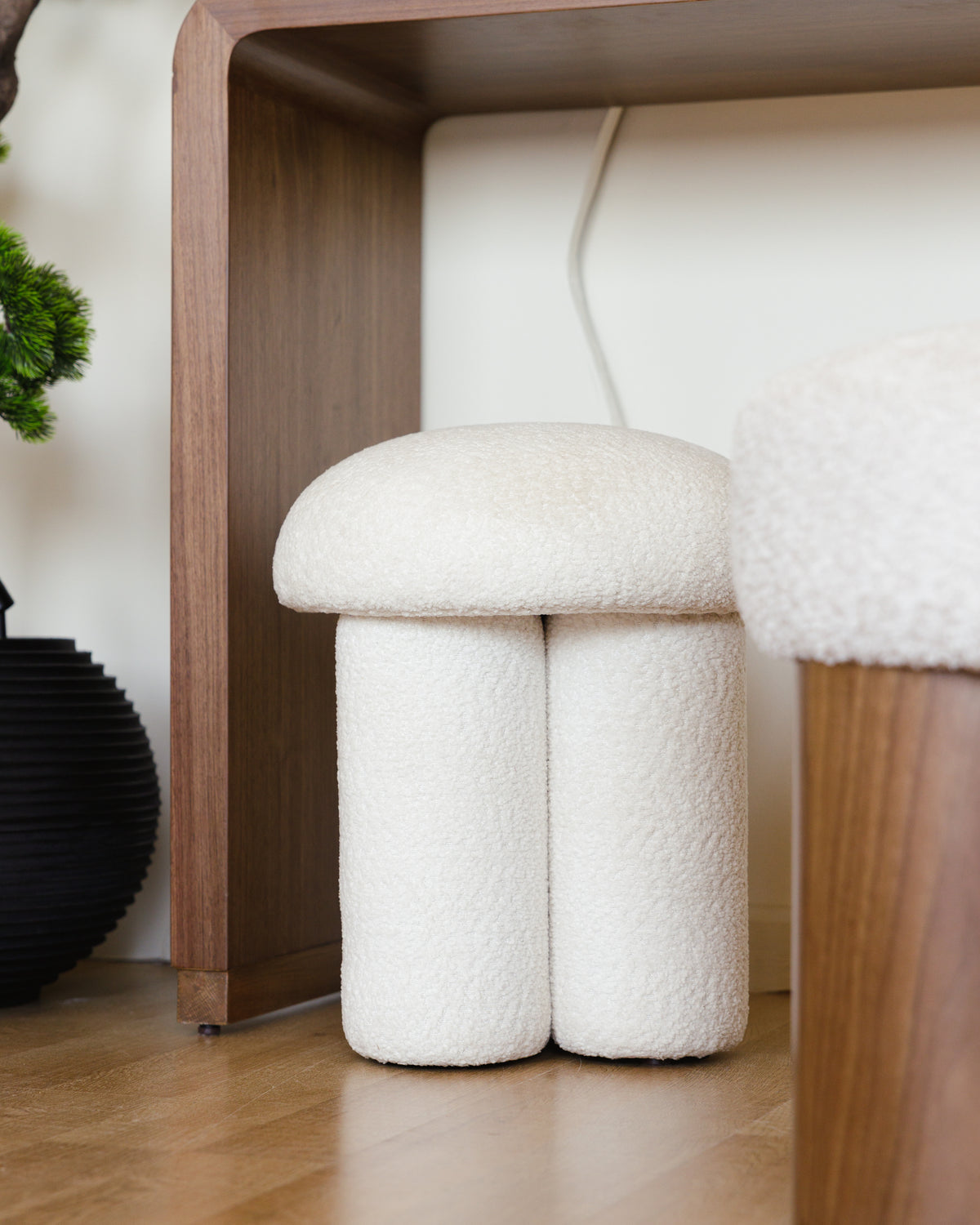 A soft, modern pouf with a dual cylindrical base and a plush, domed top is placed next to a wooden shelf. The pouf is covered in luxurious off-white bouclé, adding a cozy.