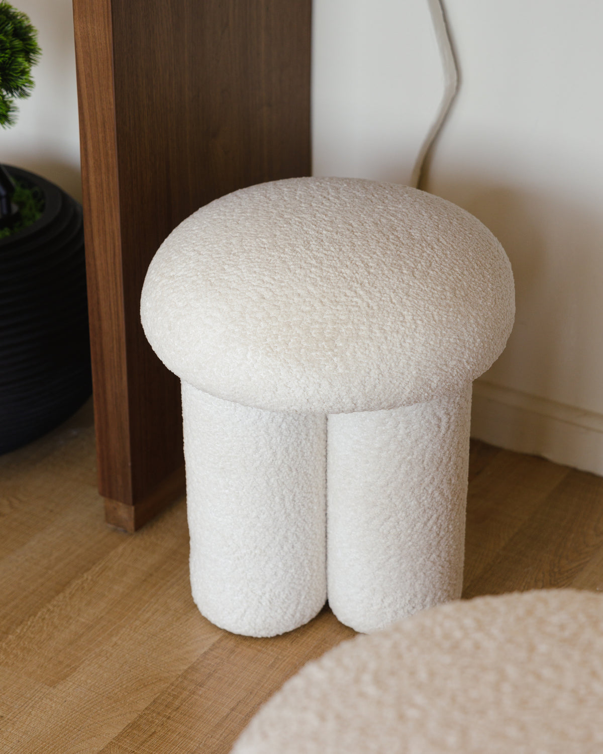 A soft, off-white bouclé pouf features a dual cylindrical base with a plush, rounded top, exuding a modern and cozy aesthetic. It is positioned next to a wooden surface, enhancing the stylish decor of the room.