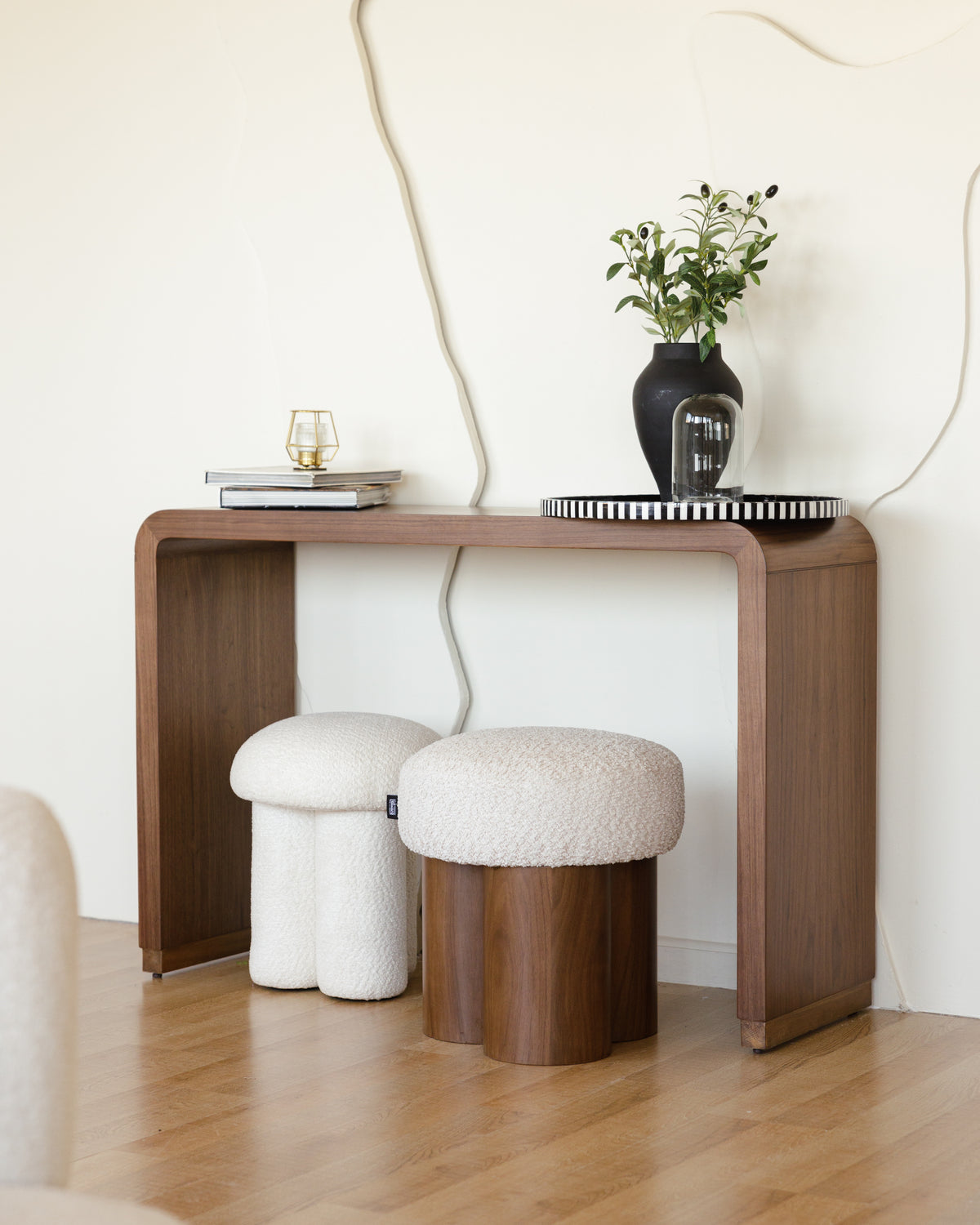 A stylish walnut console table with smooth, rounded edges stands against a light wall, accompanied by two textured stools. On the table, a small vase with greenery and a decorative tray enhance the modern aesthetic of the space.