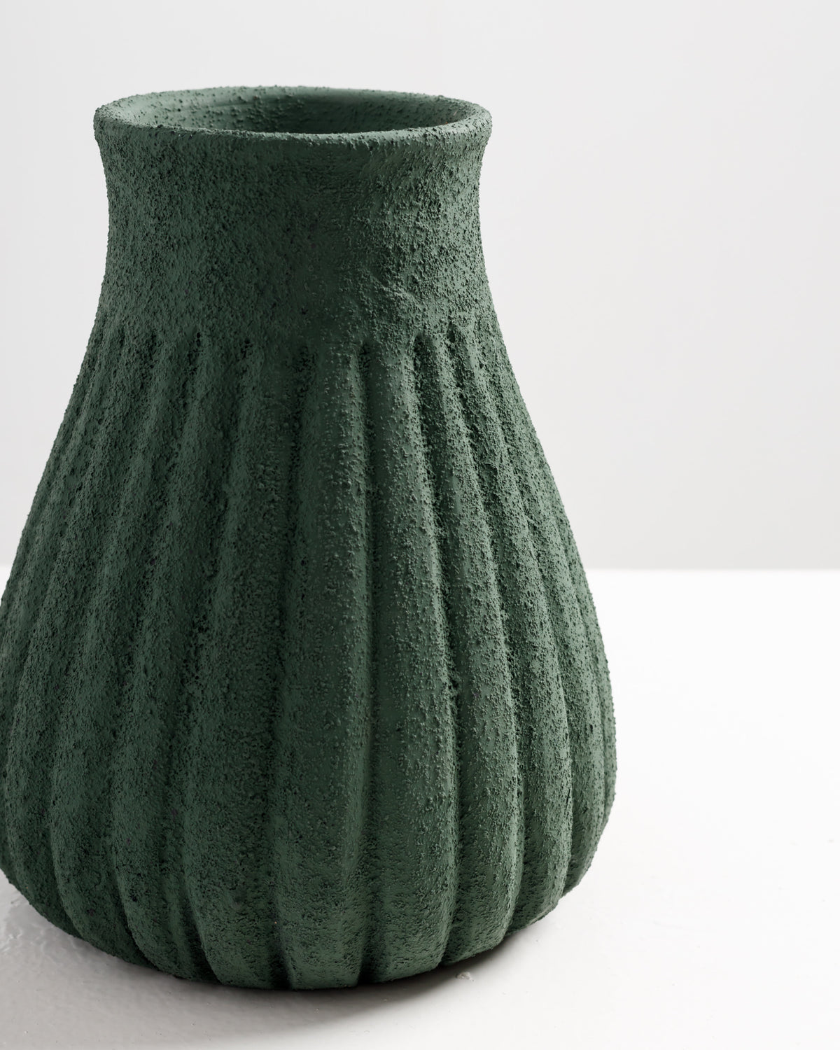 A textured, green vase with vertical ribbed patterns sits on a white surface. The vase has a rounded base that tapers towards the top, showcasing a rustic.