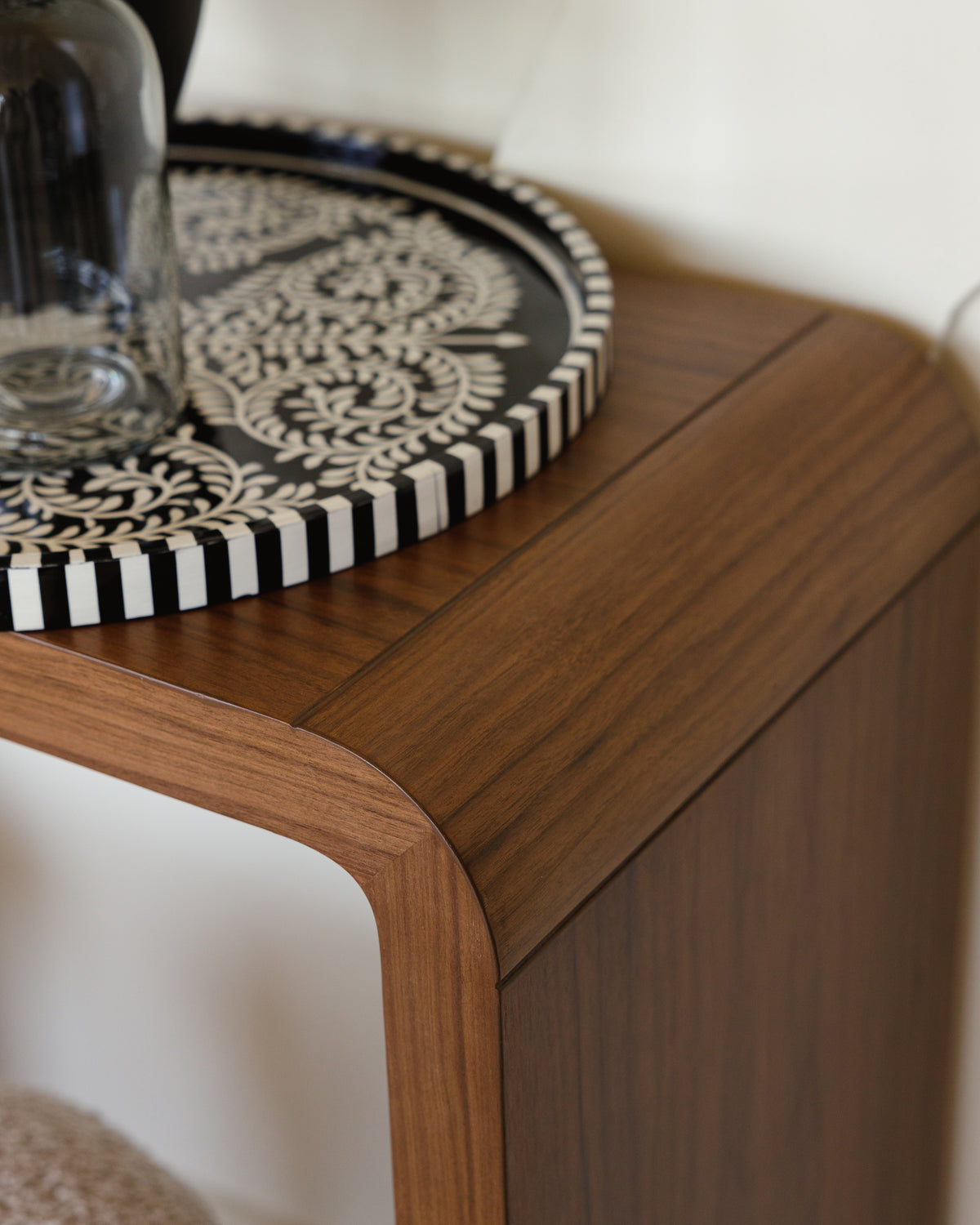 A close-up view of a walnut wood console table featuring smooth, rounded edges. The table displays a decorative black and white tray on top, highlighting its rich wood grain and elegant design.