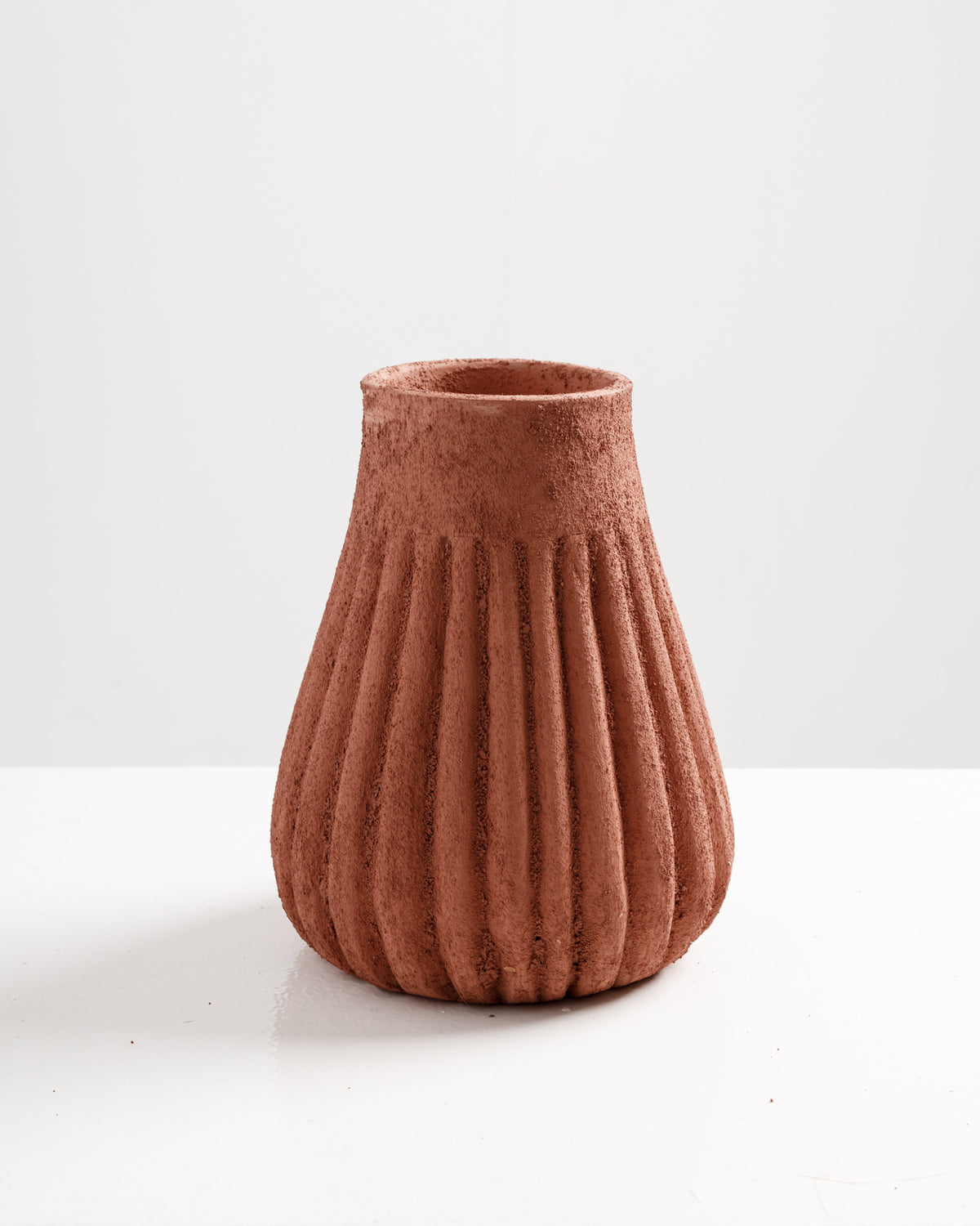 A textured terracotta vase with a fluted design sits on a white surface. The vase features a wide base that narrows towards the top, showcasing its earthy color and unique shape against a plain background.