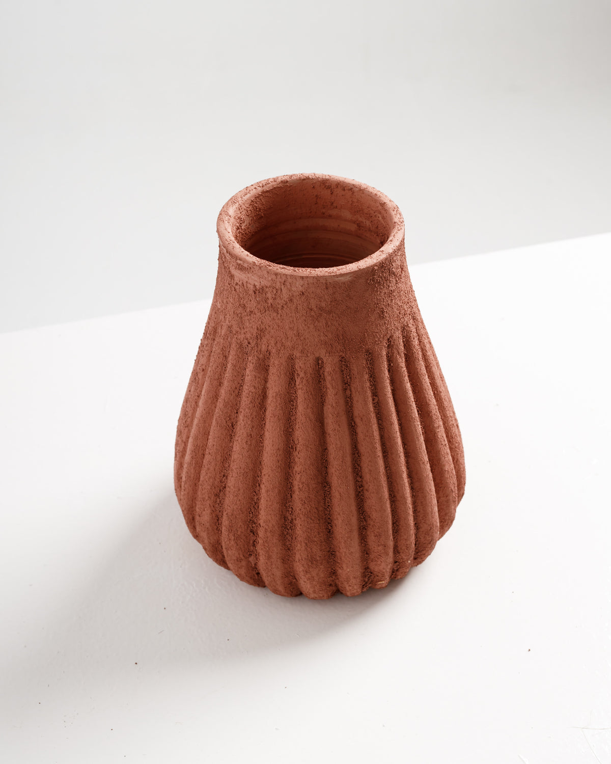 A textured terracotta vase with a fluted design sits on a white surface. The warm, earthy tones and unique shape create an elegant focal point, perfect for showcasing floral arrangements or as a standalone decorative piece.