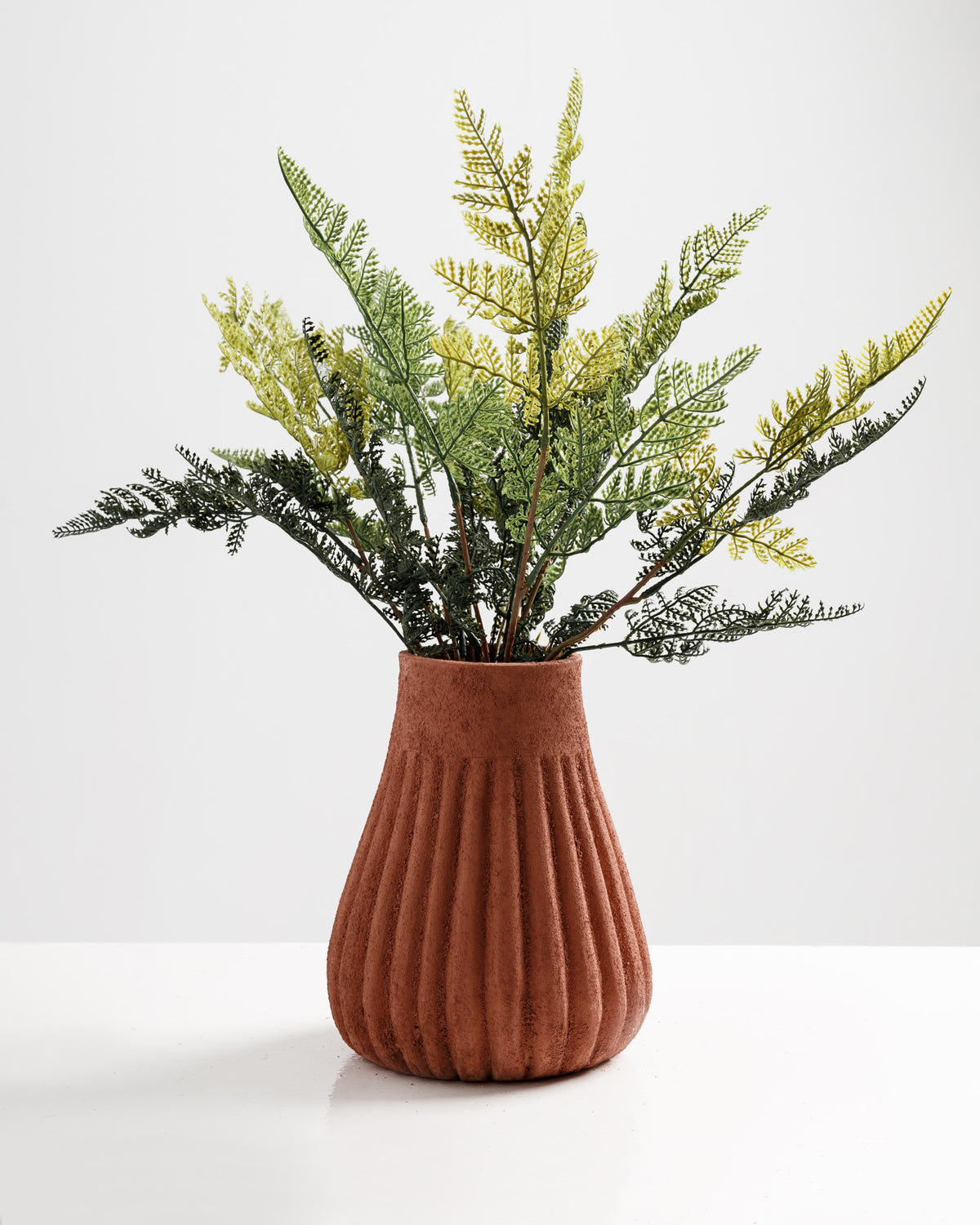 A textured terracotta vase features a fluted design and holds an arrangement of green and yellow faux foliage. The background is plain white, enhancing the vibrant colors of the decorative greenery.