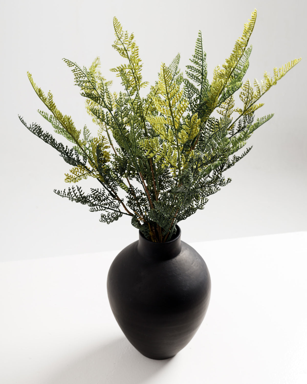 A black ceramic vase holds an arrangement of vibrant green and yellow artificial foliage. The delicate leaves are intricately designed, adding a touch of nature to the minimalist setting against a light background.