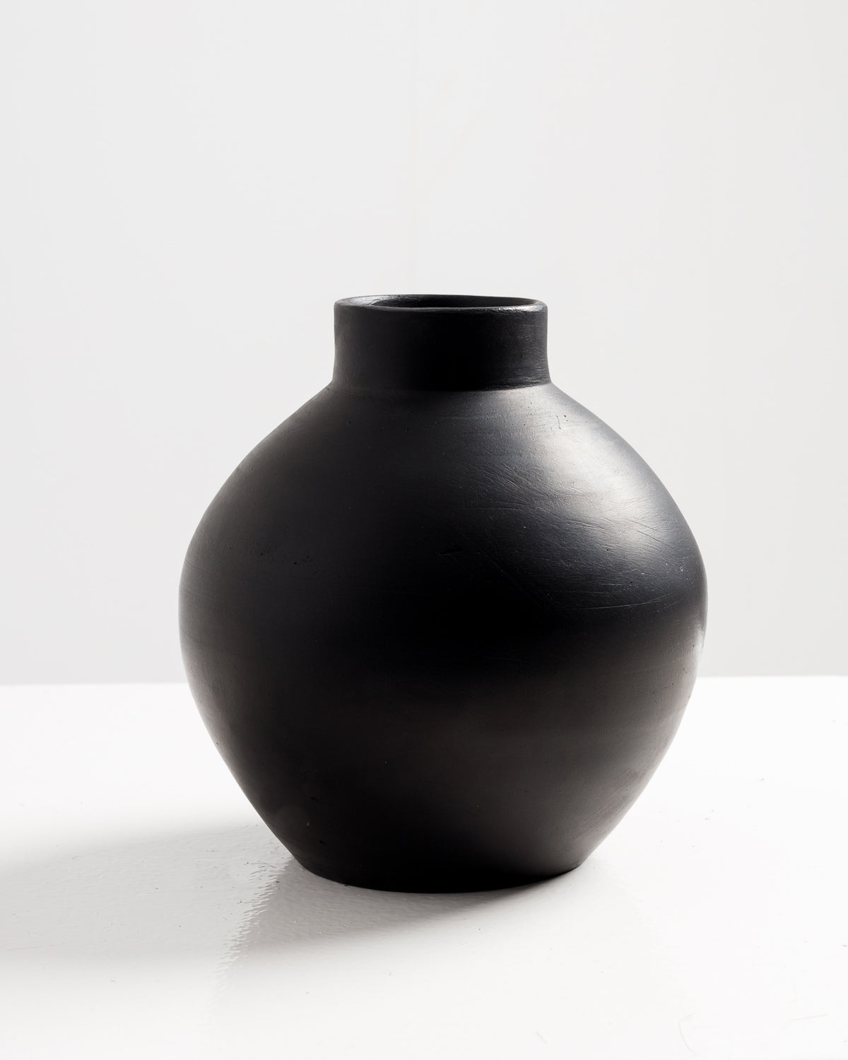 A round, matte black vase with a smooth texture and a wide body that narrows at the neck is displayed against a light background. The vase’s simple.