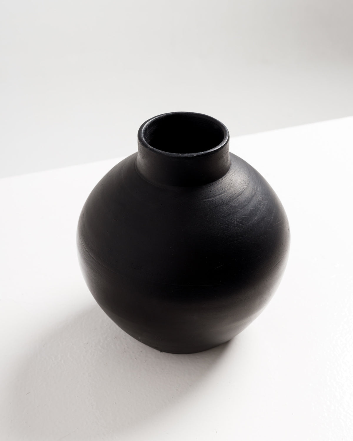 A round, matte black vase with a smooth surface and a slightly flared neck sits on a white table. The vases simple, modern design adds an elegant touch to any decor setting.