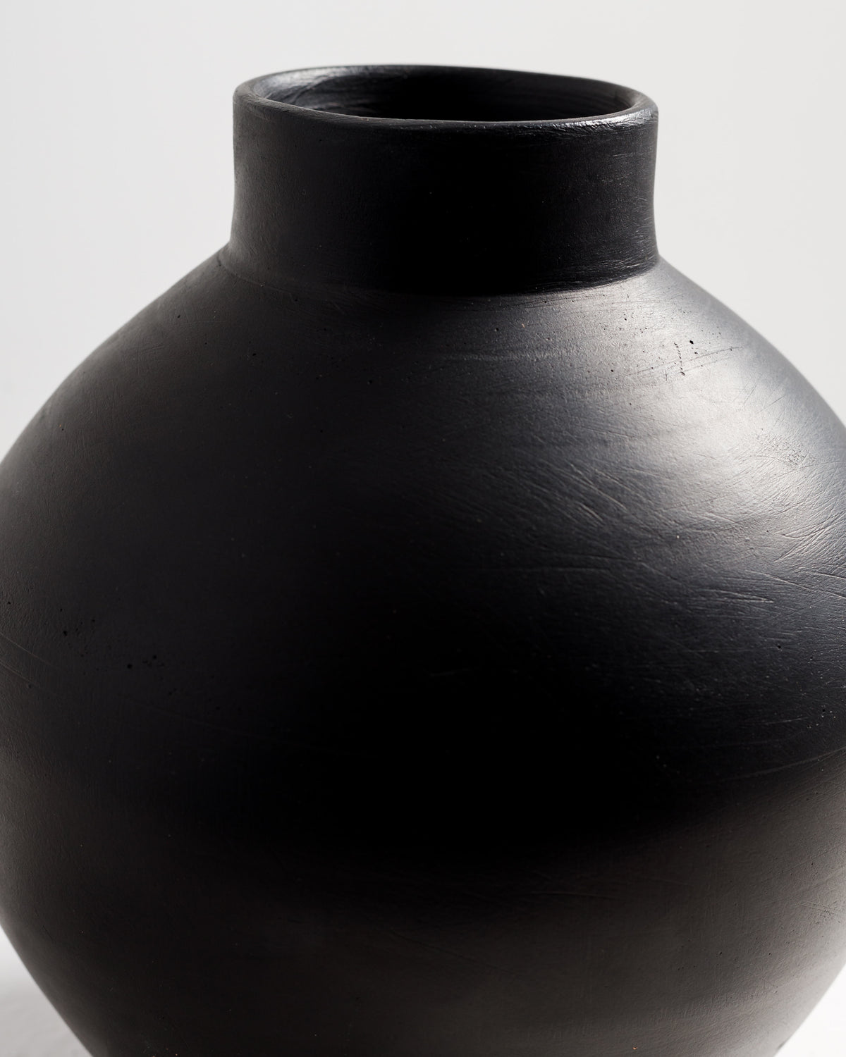 A close-up view of a round, matte black vase with a smooth surface and a narrow neck. The vase has a simple, elegant design.