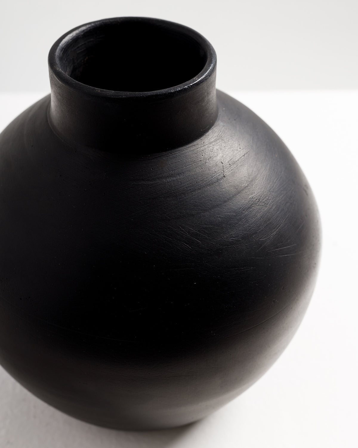 A close-up view of a round black vase with a smooth, matte finish. The vase features a wide body that tapers to a narrow neck, placed against a light-colored background for contrast.