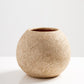 A textured, round vase with a sandy beige color sits on a white surface. The vase has a rustic appearance, featuring a slightly uneven surface that adds to its organic charm, against a simple, light backdrop.