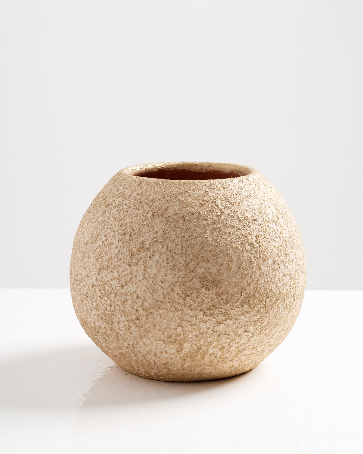A textured, round vase with a sandy beige color sits on a white surface. The vase has a rustic appearance, featuring a slightly uneven surface that adds to its organic charm, against a simple, light backdrop.