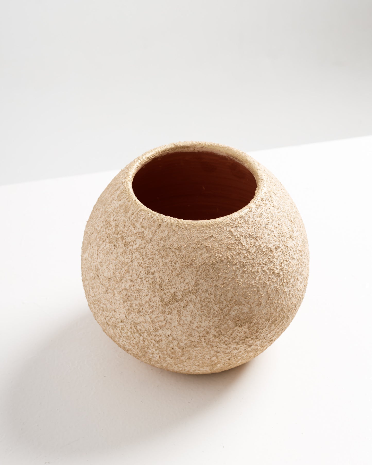 A textured, round vase made of natural materials sits on a white surface, featuring a warm, earthy tone. The vase has a wide opening at the top, emphasizing its organic shape and rustic charm.
