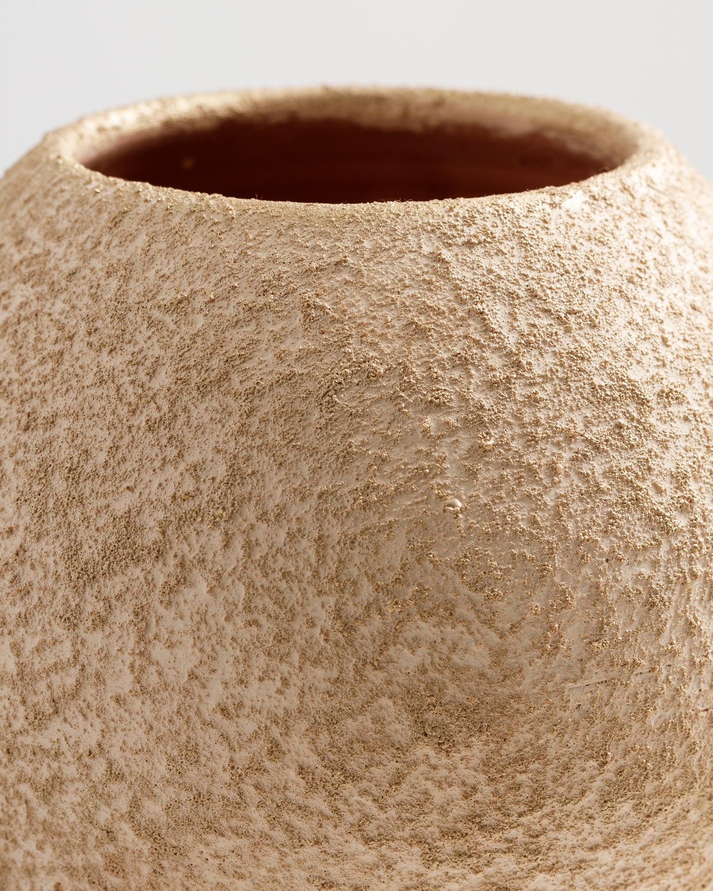 Close-up view of a textured, beige vase featuring a subtle gold sheen. The surface appears rough and organic, highlighting the craftsmanship and unique finish of the piece.
