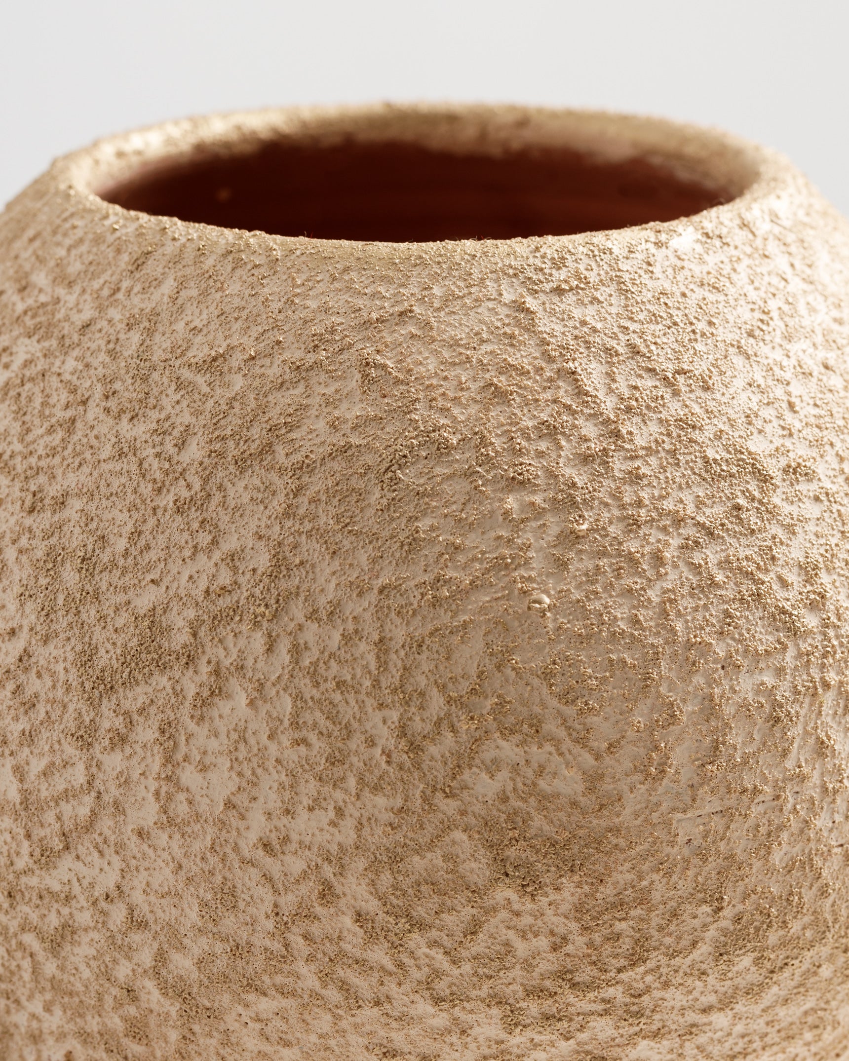 Close-up view of a textured, beige vase featuring a subtle gold sheen. The surface appears rough and organic, highlighting the craftsmanship and unique finish of the piece.
