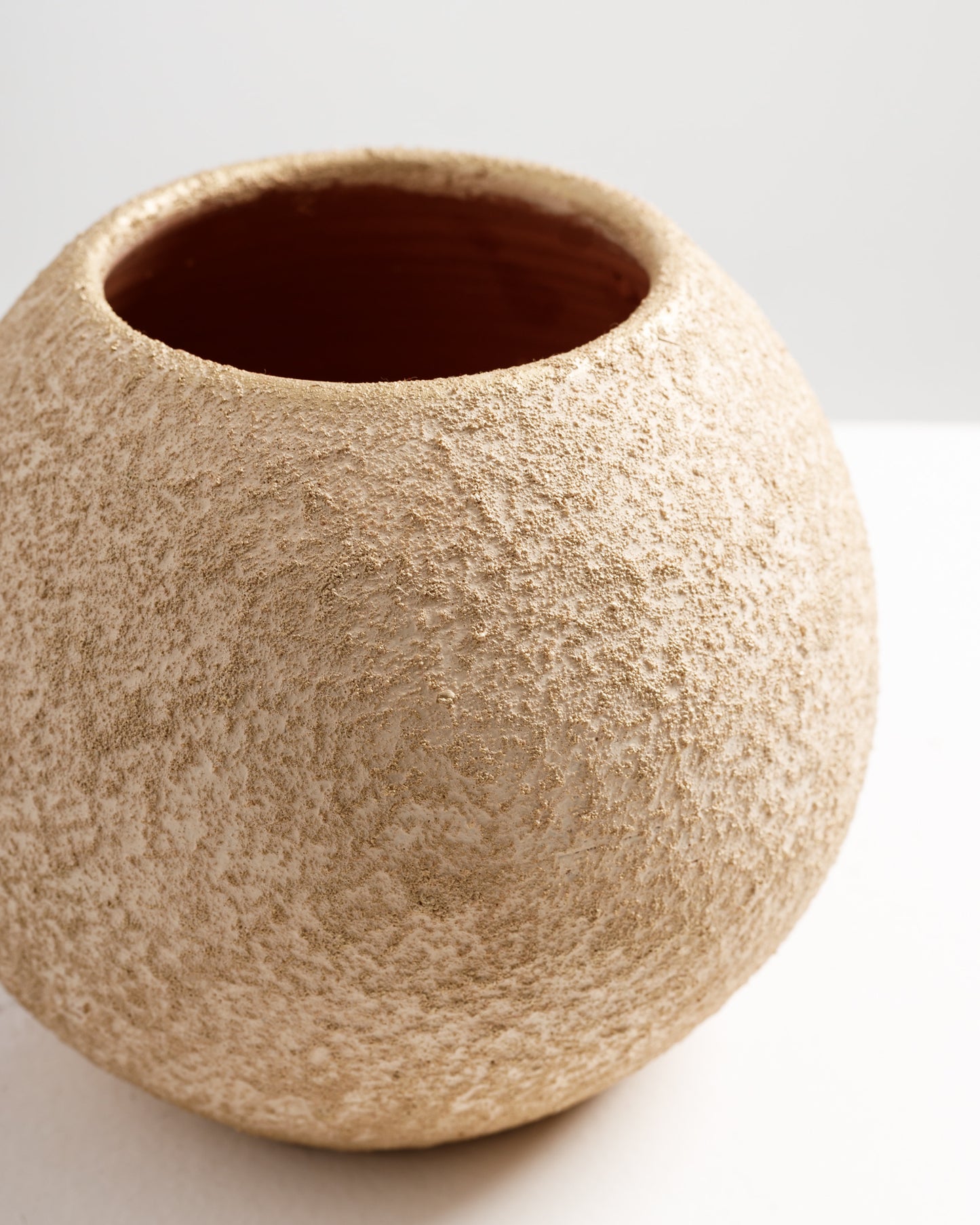 A close-up view of a textured, round gold vase with a rough, matte finish. The interior of the vase is a deep reddish-brown, contrasting with the shimmering gold exterior.