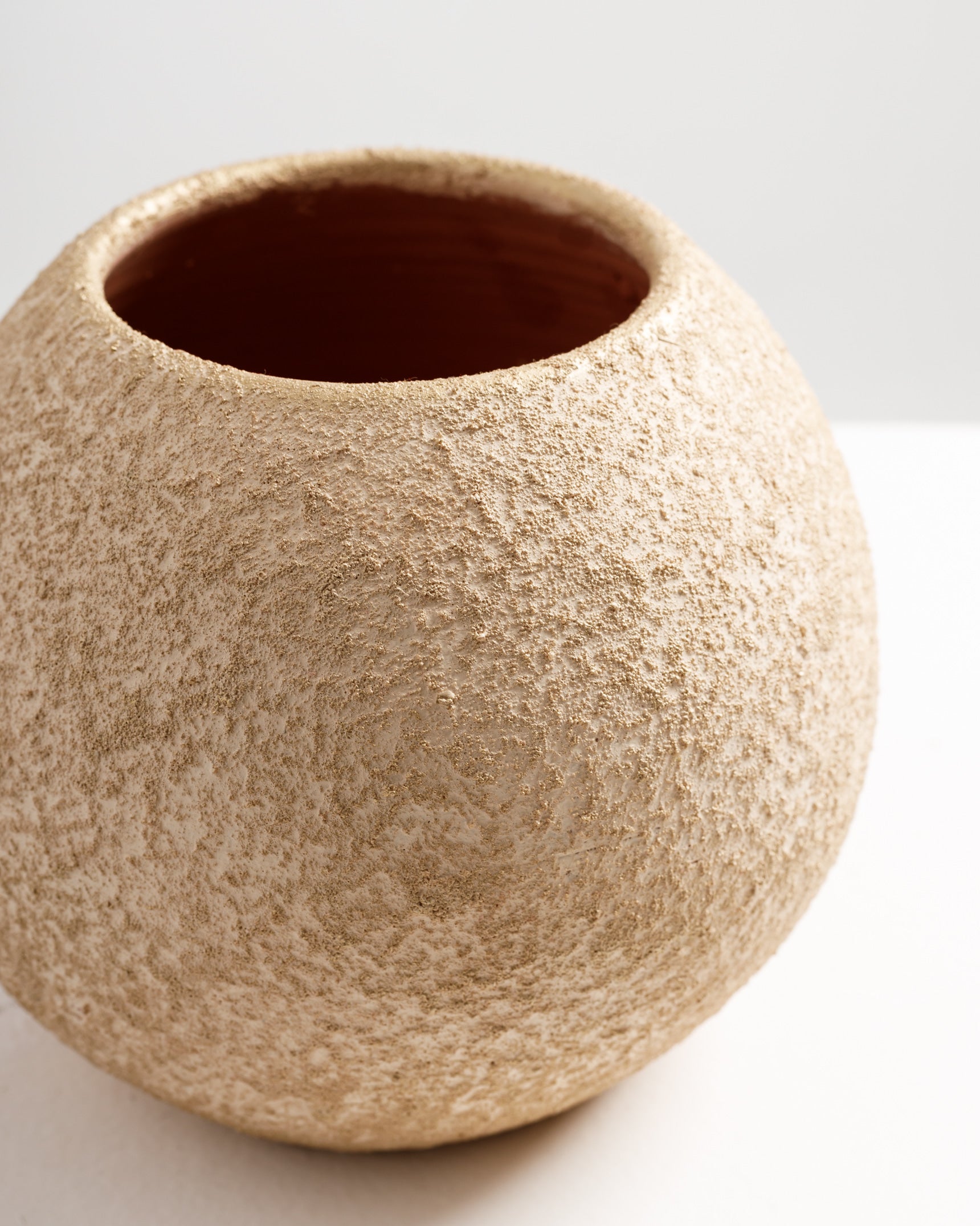 A close-up view of a textured, round gold vase with a rough, matte finish. The interior of the vase is a deep reddish-brown, contrasting with the shimmering gold exterior.
