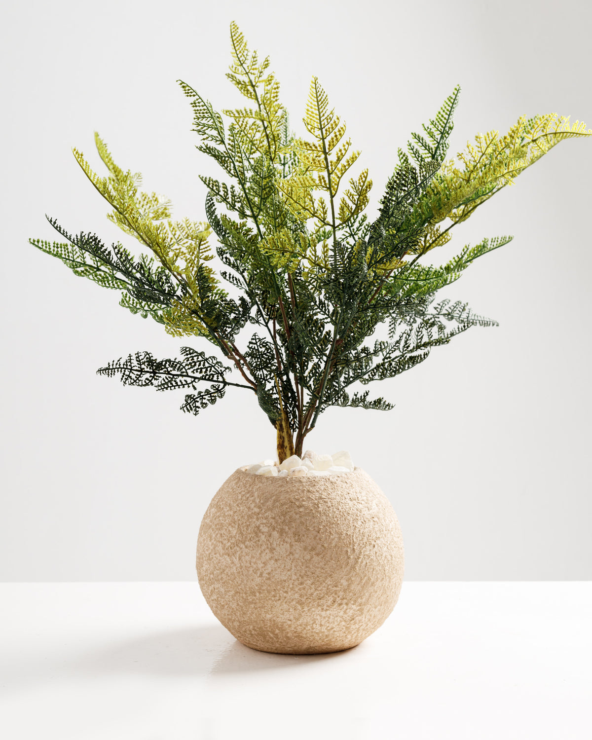 A decorative faux plant featuring vibrant green leaves is displayed in a textured, round beige pot. The arrangement adds a touch of greenery and elegance to any interior space.