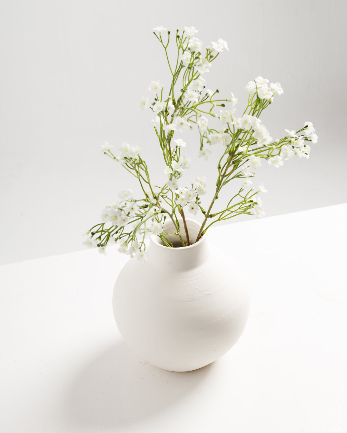A minimalist white vase holds delicate white flower branches, showcasing small blooms and slender green stems. The vase is positioned on a light surface against a soft, neutral background, enhancing the serene aesthetic.