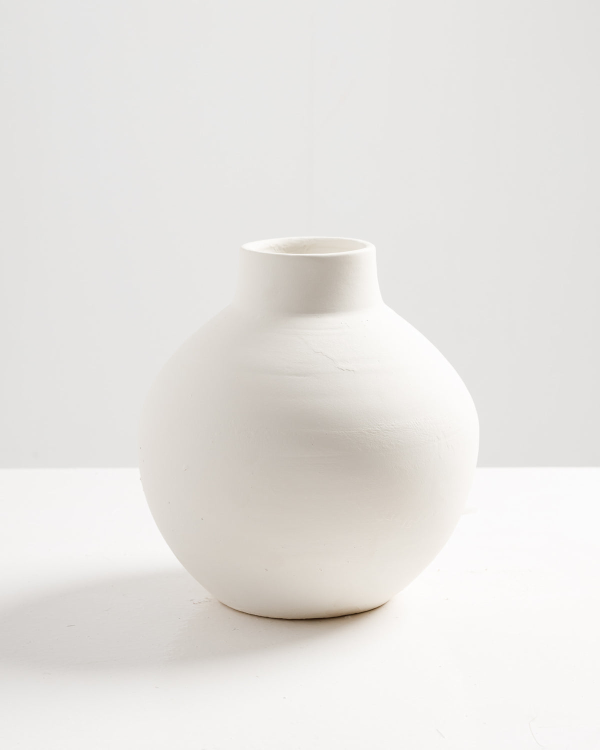 A minimalist round vase made of smooth, unglazed white ceramic is displayed against a plain white background. The vase features a rounded body with a narrow neck, emphasizing its contemporary design.