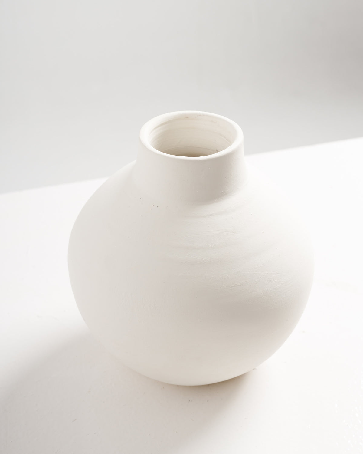 A simple, round white vase with a smooth, matte finish sits on a light surface. The vase features a gently curved body and a narrow opening at the top, creating an elegant and minimalist design.