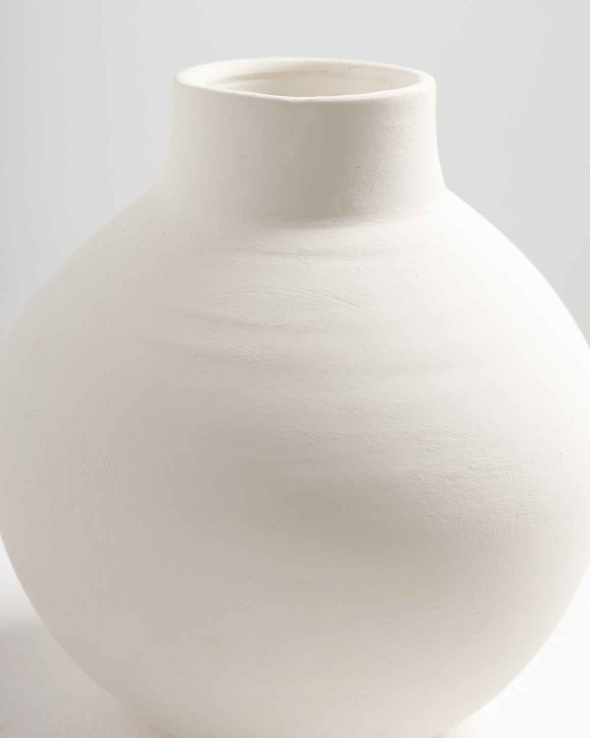 A smooth, round white vase with a narrow neck and a slightly textured surface is displayed against a light background. The vases minimalist design highlights its elegant, contemporary shape.