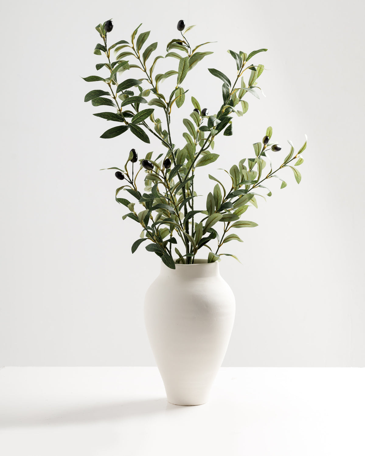 A stylish white vase holds an elegant arrangement of artificial olive branches featuring green leaves and black foam fruits. The minimalist design.