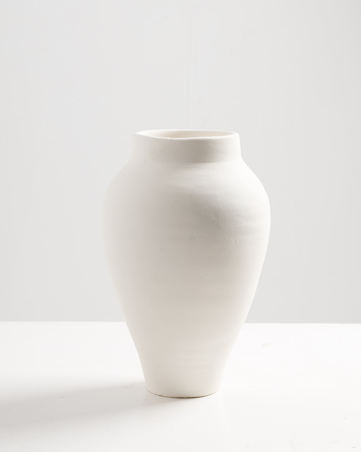 A tall, elegant white ceramic vase with a smooth, matte finish sits on a white surface. The vase features a rounded body that tapers at the base, creating a simple yet sophisticated design against a minimalistic background.