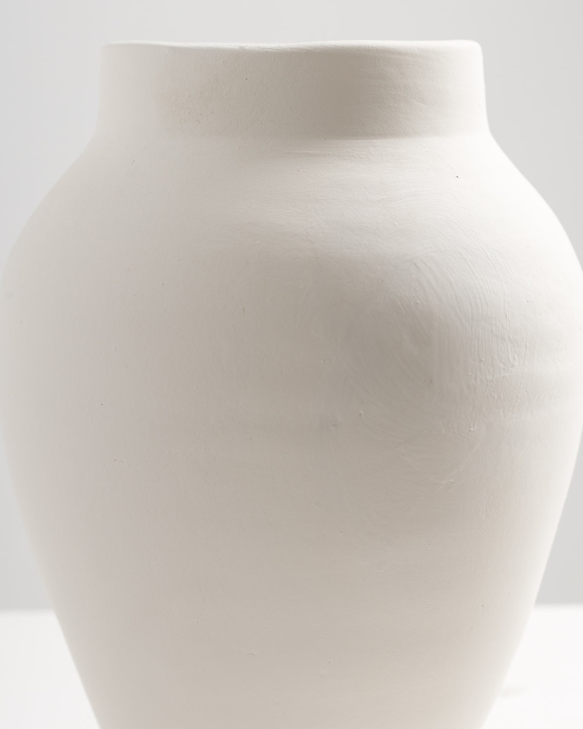 A smooth, white ceramic vase with a rounded body and a wide mouth, set against a minimalistic background. The vase has a matte finish, emphasizing its elegant, simple design.