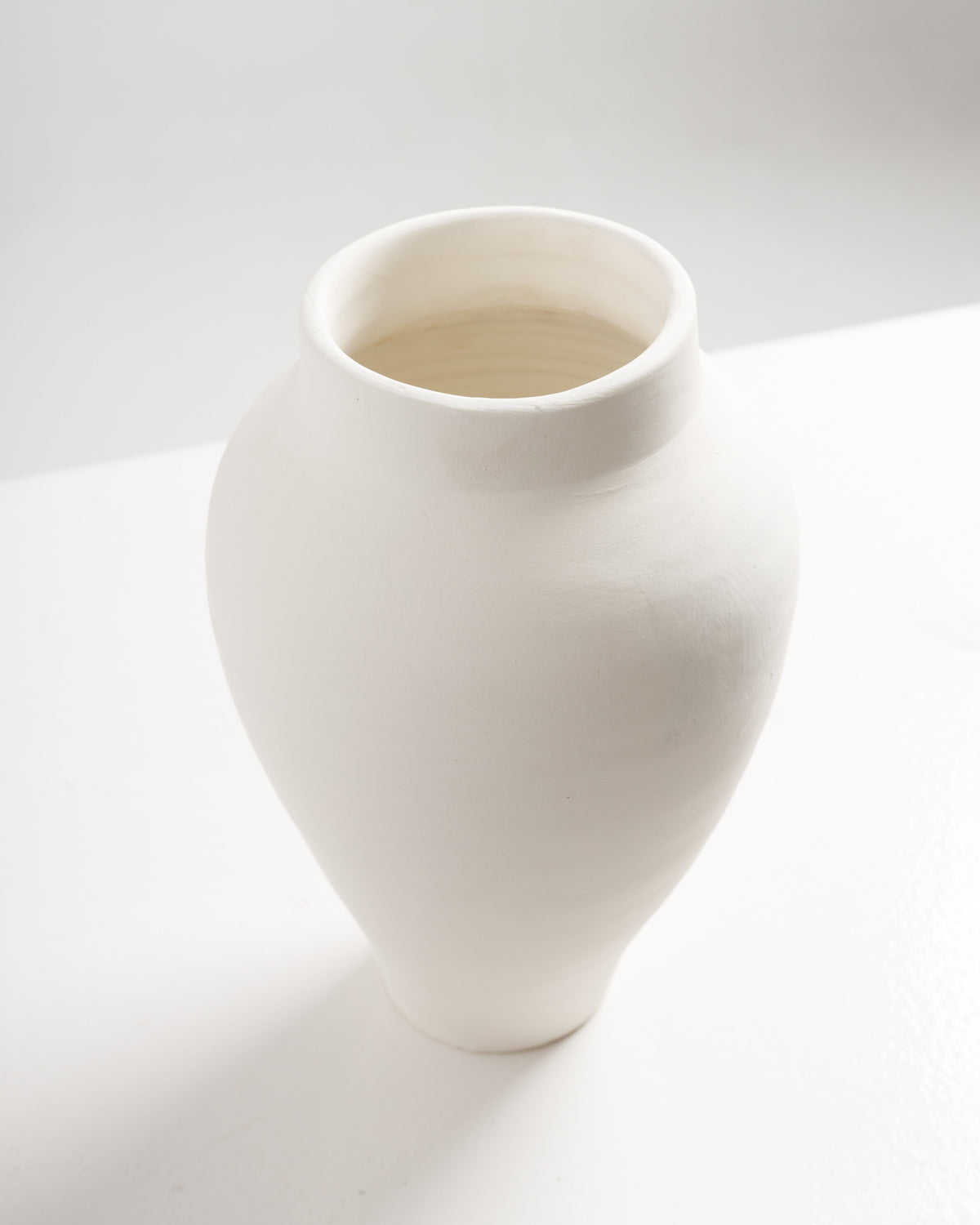A smooth, unglazed white ceramic vase with a rounded body and narrow neck sits on a light-colored surface. Its minimalist design showcases the natural texture of the clay, making it a versatile decorative piece.