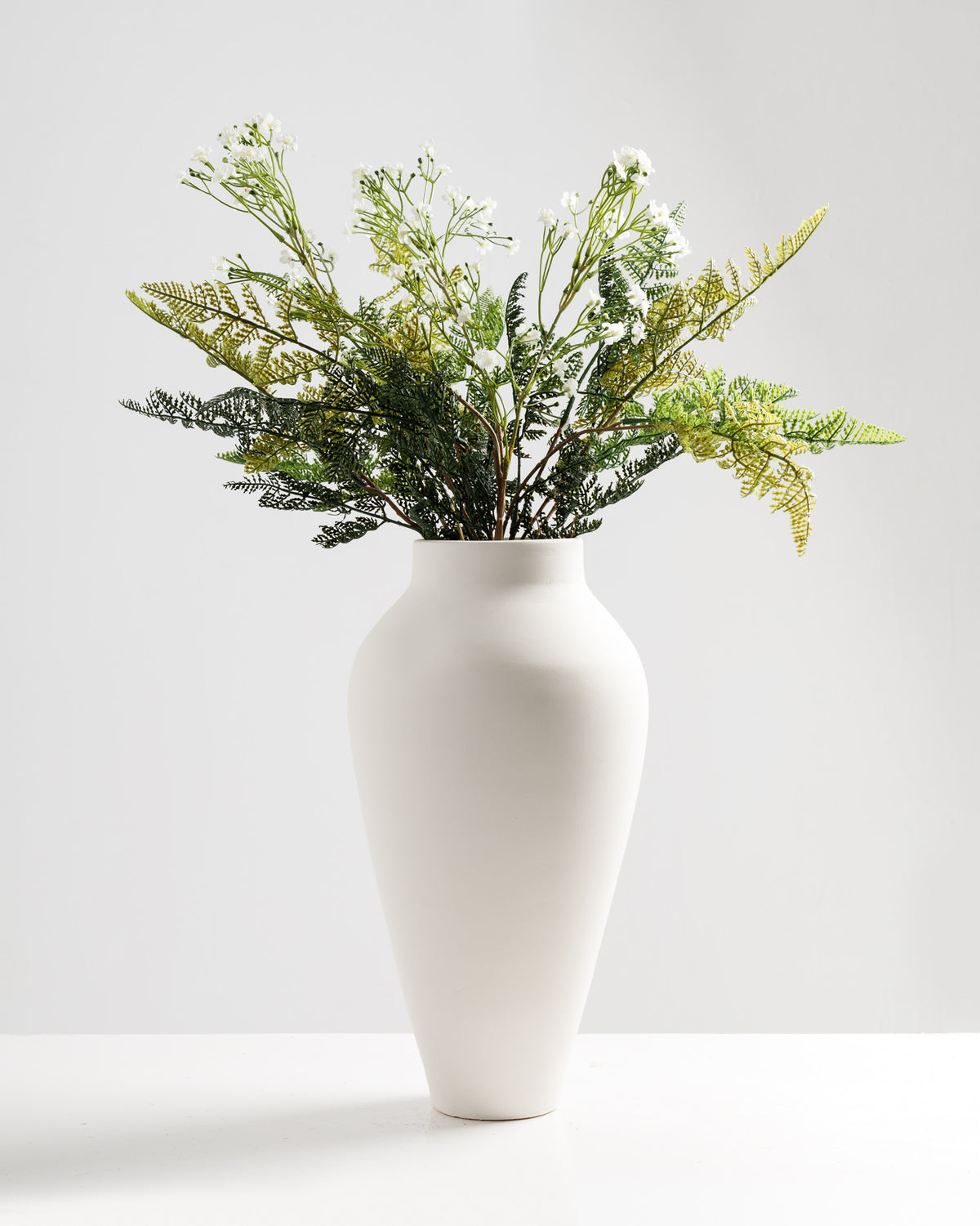 A tall, elegant white vase features a sleek, rounded shape and is filled with an arrangement of delicate white flowers and lush green ferns.