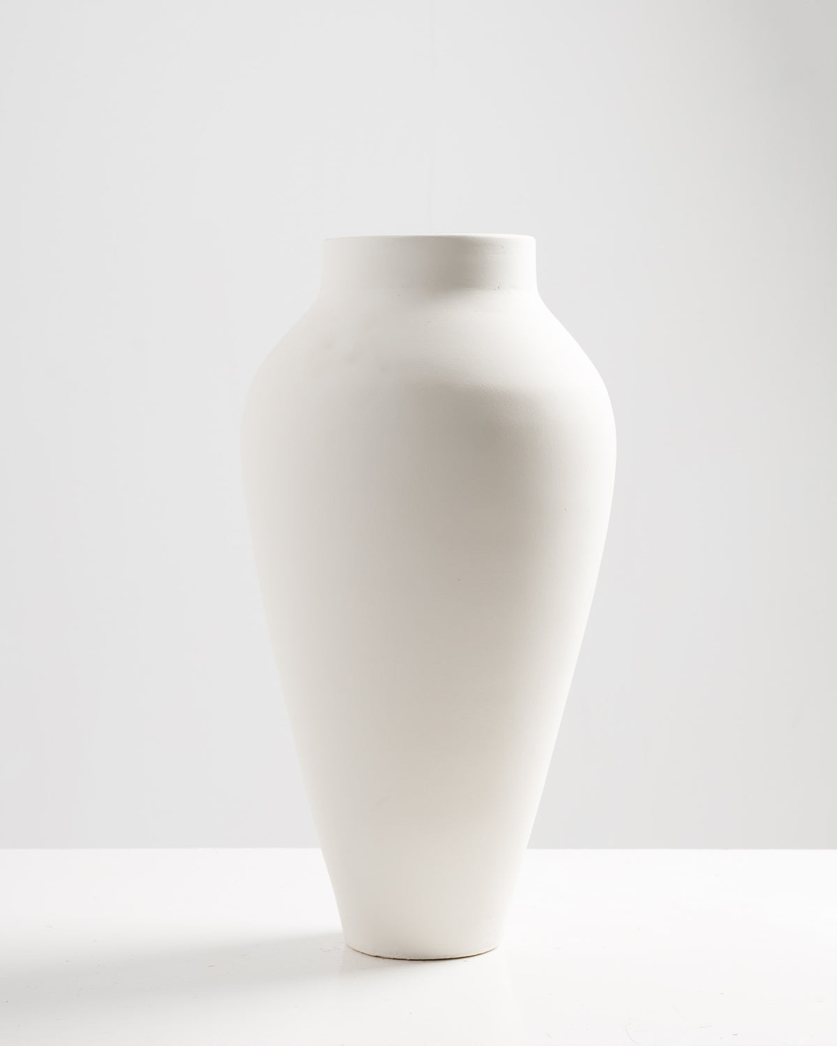 A tall, elegant white vase with a smooth, rounded body and a narrow neck stands against a soft, light background. The minimalist design emphasizes its modern aesthetic.